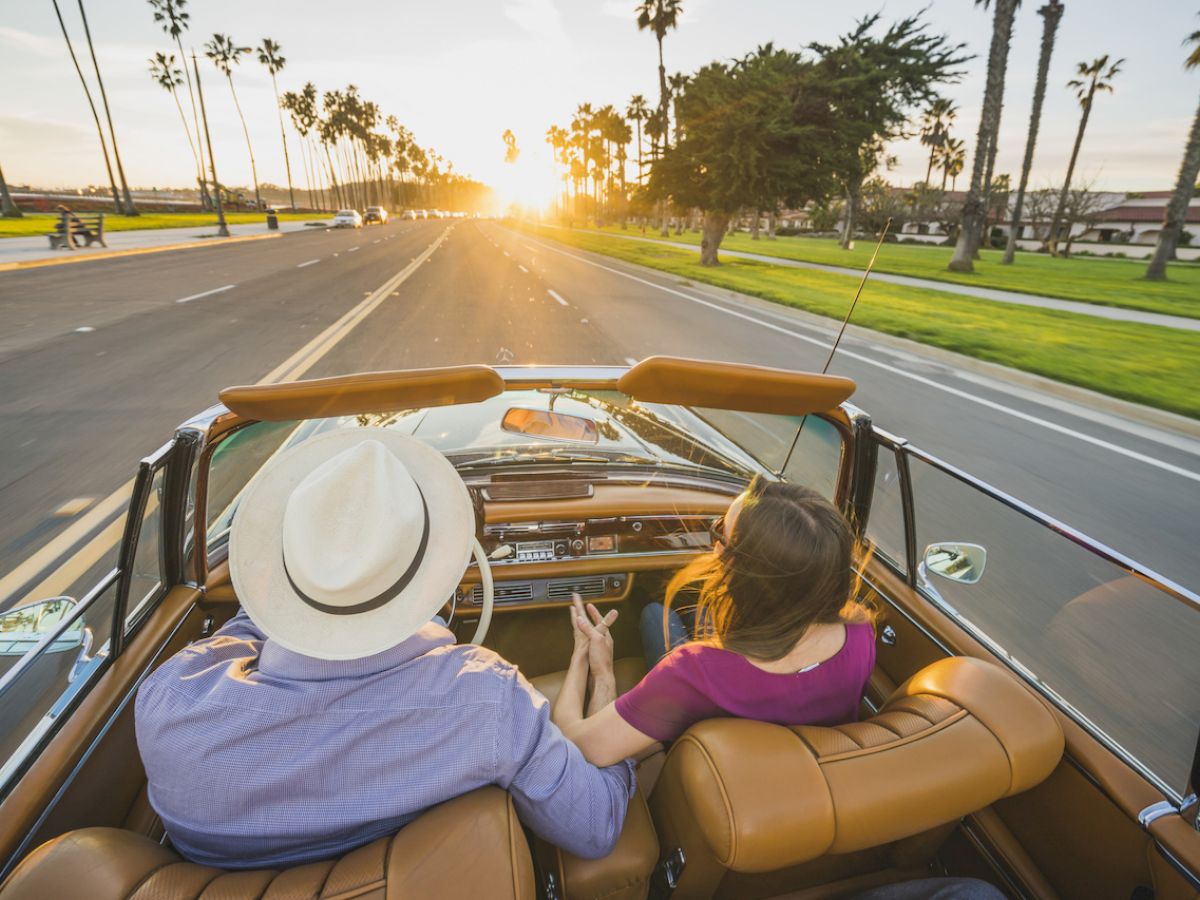 A couple is driving a classic convertible down a scenic road at sunset, holding hands. Palm trees line the sides of the road.