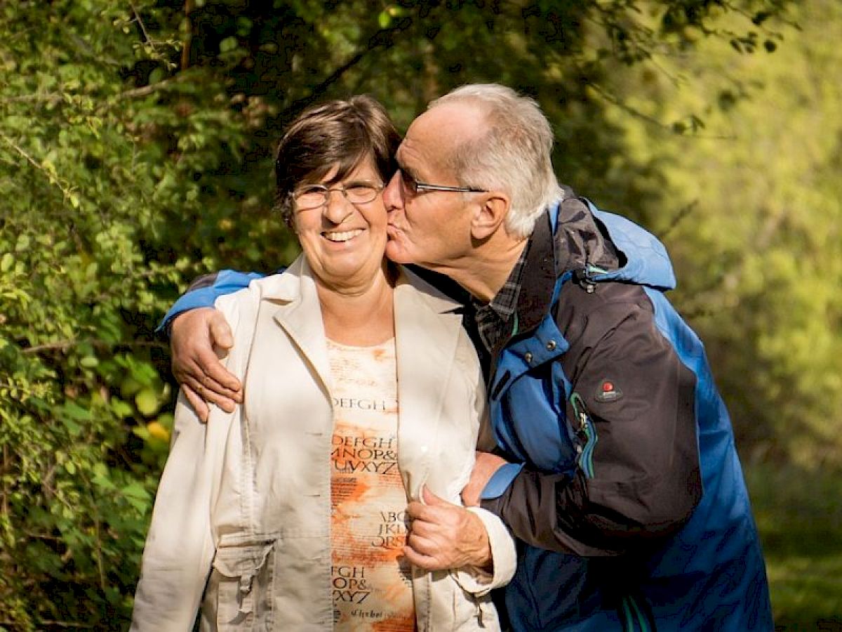 An older man kisses a smiling woman on the cheek while walking together outdoors, surrounded by greenery, with sunlight filtering through the trees.