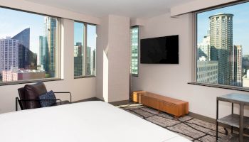 A modern hotel room with large windows showcasing a cityscape, a wall-mounted TV, and a bed with a chair near the window completes the scene.