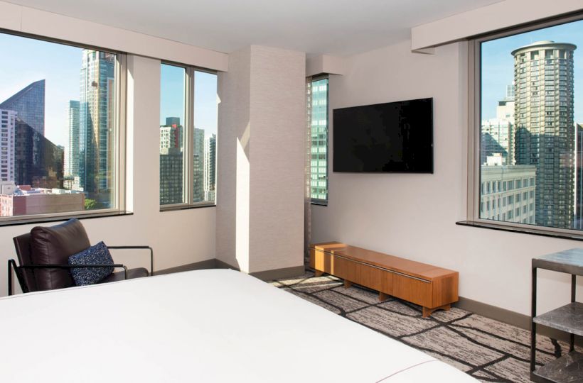 A modern hotel room with large windows showcasing a cityscape, a wall-mounted TV, and a bed with a chair near the window completes the scene.