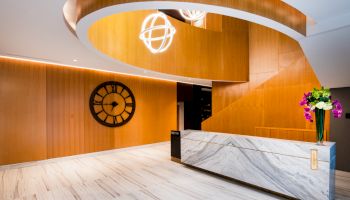 A modern lobby features a clock on the wall, a marble reception desk, and a spiral staircase with two globe chandeliers overhead.