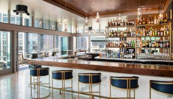 A modern bar with high shelves filled with various bottles, four bar stools, large windows, and stylish lighting fixtures.