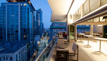The image shows a modern, high-rise building's balcony overlooking a city street at dusk, with tables and chairs set up for outdoor seating.