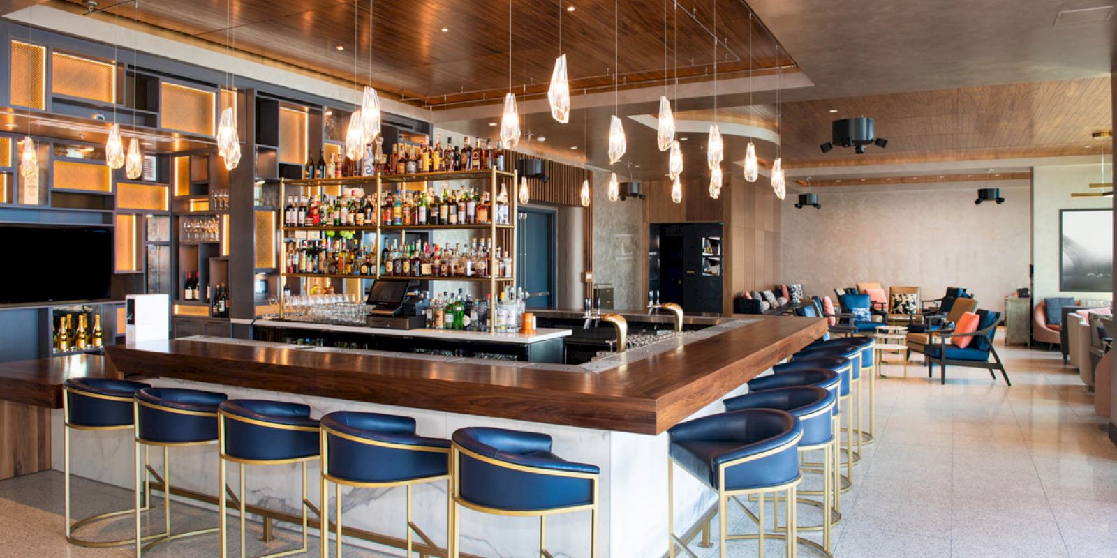The image shows a modern bar with wooden accents, hanging light fixtures, and blue cushioned barstools. There are bottles of various beverages displayed on shelves.