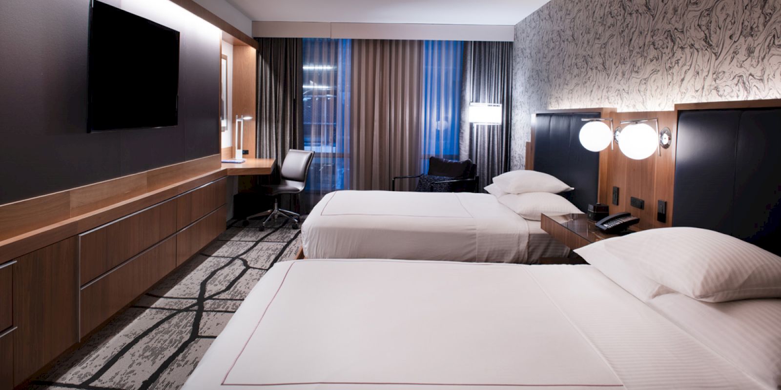 The image shows a modern hotel room with two neatly made beds, a wall-mounted TV, a desk, a chair, and contemporary lighting fixtures.