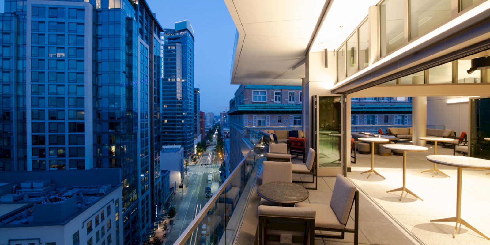 An elegant balcony overlooks a cityscape at dusk, featuring modern furniture, well-lit tables, and floor-to-ceiling windows.