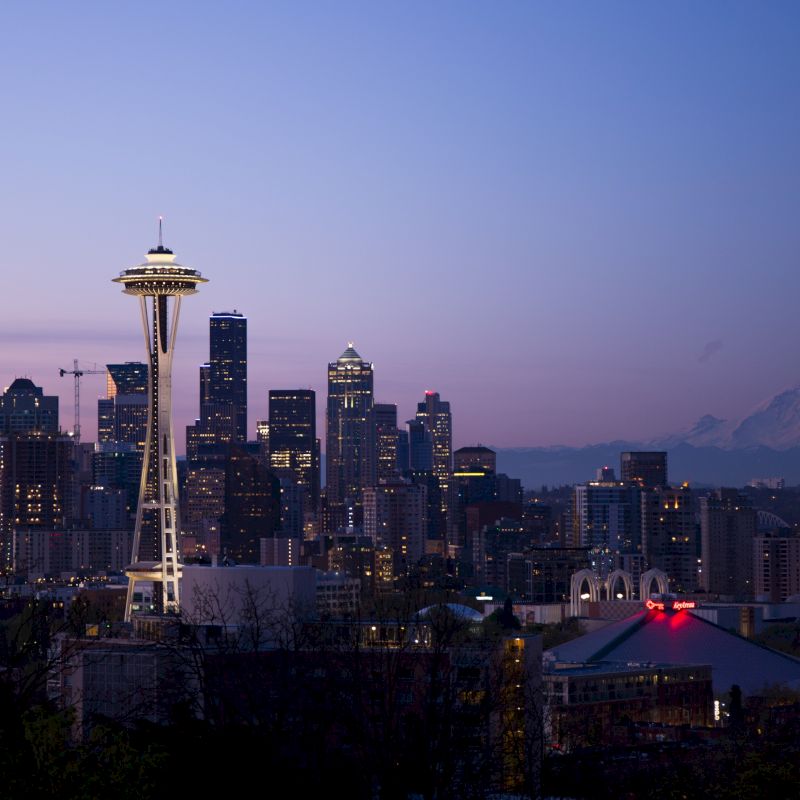 A cityscape featuring a tall tower, assorted skyscrapers, and a distant mountain under a twilight sky, likely taken at dusk.