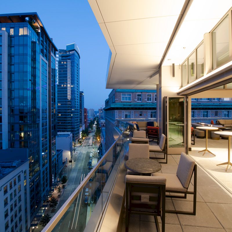 A modern balcony with tables and chairs overlooks a city street lined with tall buildings, with a warm, illuminated interior visible through glass windows.