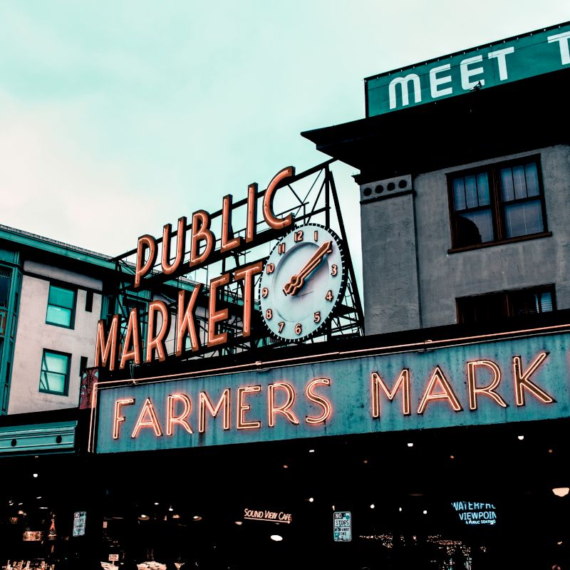 The image shows a public farmers market with a neon sign and clock, adjacent buildings, and a partial view of a "hotel" sign.
