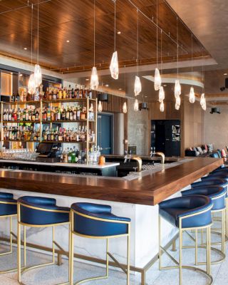 A modern bar with blue and gold stools, hanging lights, a fully stocked liquor shelf, and seating areas visible in the background.