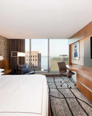 The image shows a modern hotel room with a large bed, flat-screen TV, desk, and windows offering a cityscape view, featuring modern decor.
