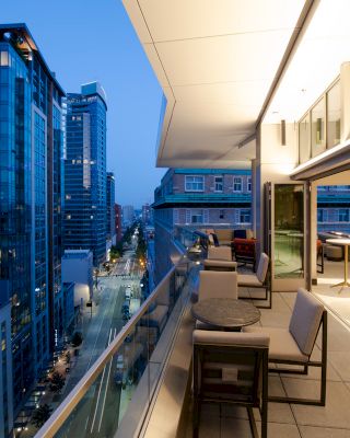 An image of a modern city view from a balcony at dusk, with outdoor seating and a spacious indoor area visible through glass doors.
