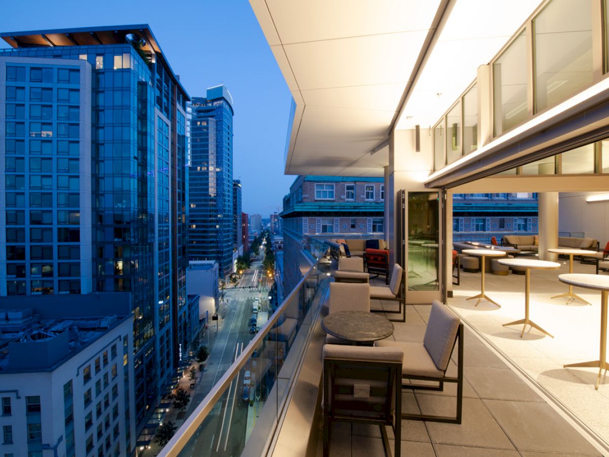 A modern cityscape at dusk features tall buildings and a stylish outdoor terrace with tables, chairs, and a glass railing overlooking the street.