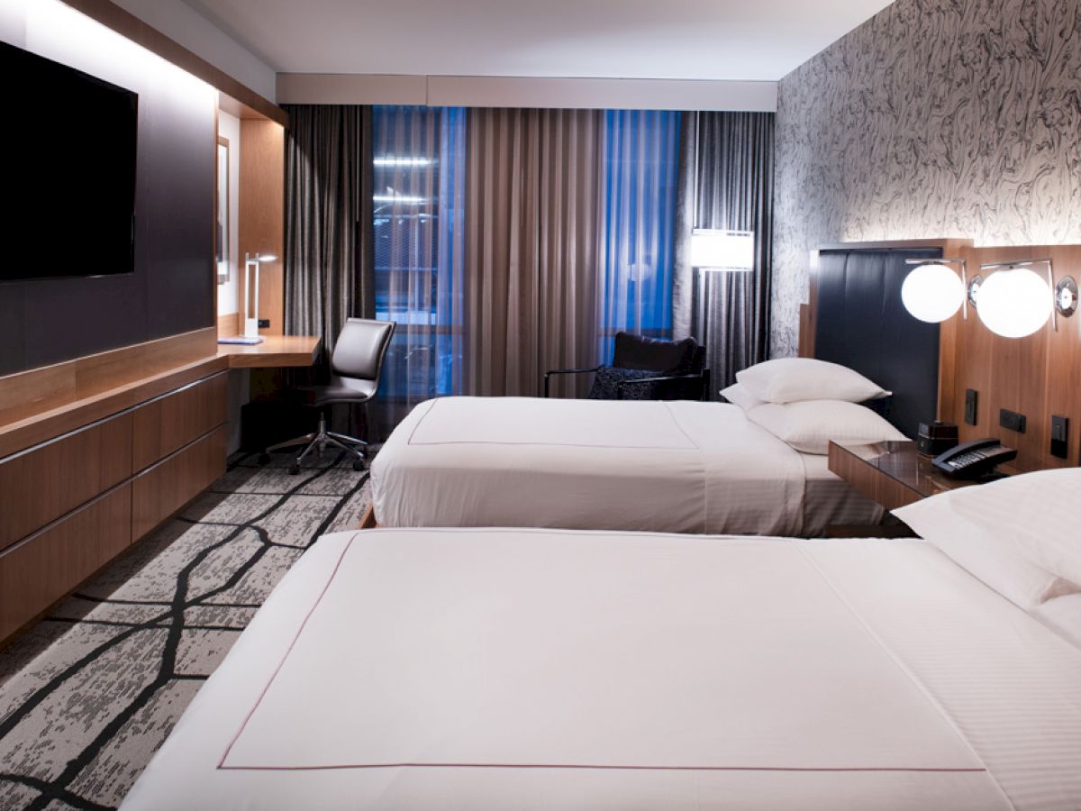 A modern hotel room with two double beds, a wall-mounted TV, a desk with a chair, stylish lighting, and floor-to-ceiling windows with curtains.