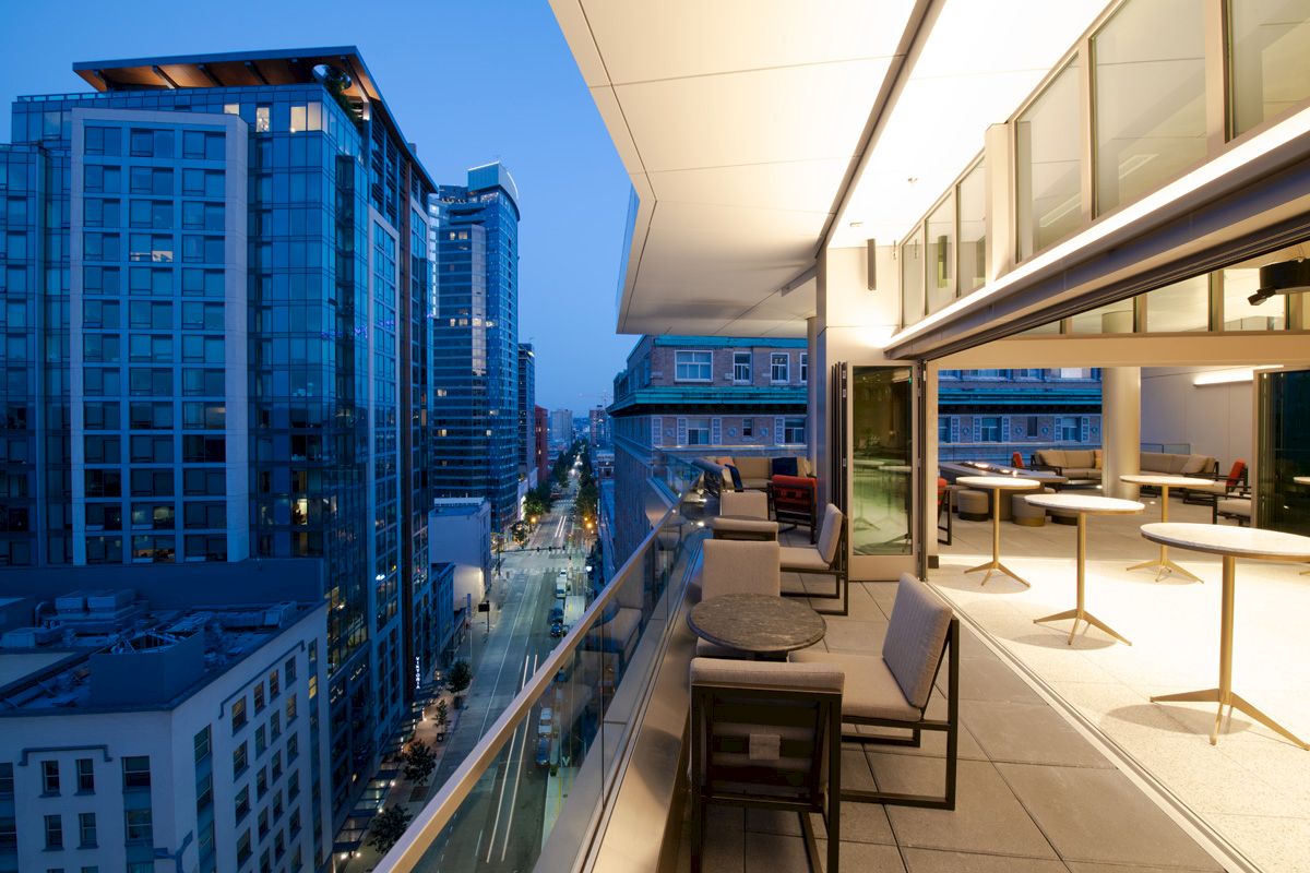 A modern balcony overlooking a city street, featuring tables, chairs, and outdoor lighting, with adjacent tall buildings illuminated at dusk.