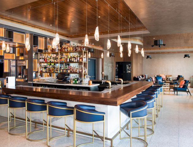 A modern bar with a large selection of liquors, stylish pendant lights, blue bar stools, and a seating area with couches in the background.