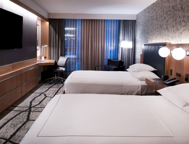 A modern hotel room with two beds, a desk and chair by the window, a TV on the wall, and bedside lamps. Room has a contemporary design.