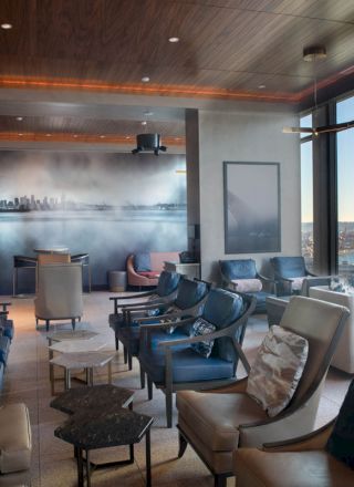 The image shows a modern lounge with comfortable seating, large windows offering views of a waterfront and Ferris wheel, and ambient lighting.