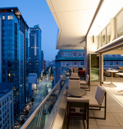 The image displays a modern cityscape viewed from a high-rise balcony with seating and city lights in the distance during dusk.
