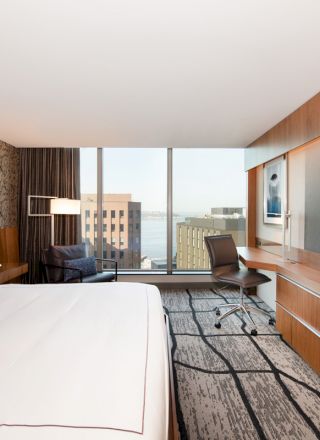 The image features a modern hotel room with a bed, a large TV, a desk and chair, and a window with a city view in the background.