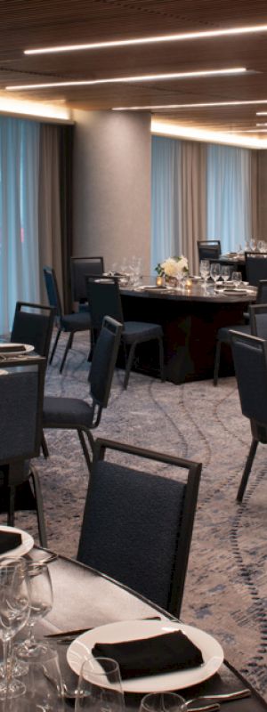 A banquet hall is set up with round tables covered in dark cloths, black chairs, and table settings, featuring minimalist floral centerpieces and candle holders.