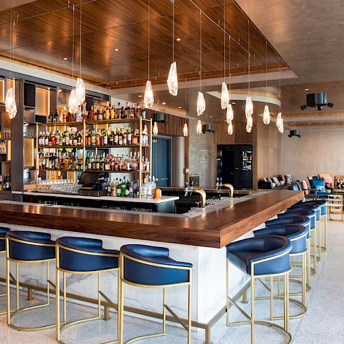 A modern bar with blue barstools, a wooden countertop, hanging lights, and a fully stocked back wall. Lounge seating is seen in the background.