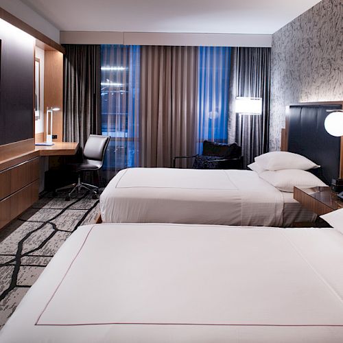 A modern hotel room with two beds, a wall-mounted TV, a desk with a chair, and contemporary lighting and decor, featuring large windows with curtains.