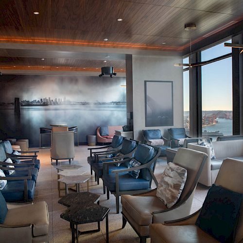 A modern lounge with ample seating, large windows offering city and water views, and a wall mural depicting a city skyline.
