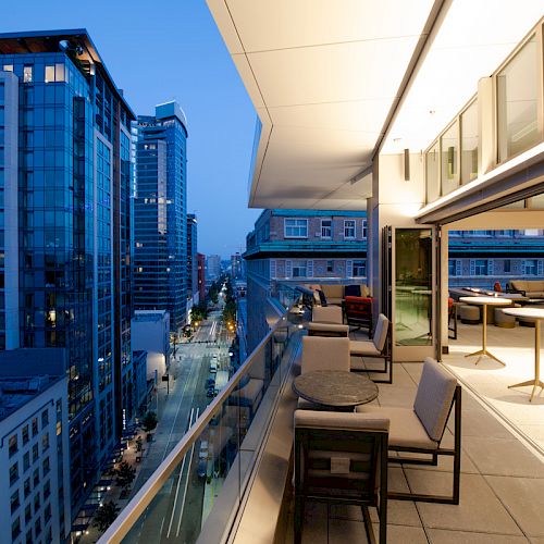 A cityscape view from a modern balcony at dusk, showing high-rise buildings and an outdoor seating area with tables and chairs.