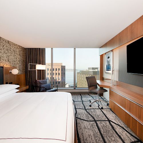 A modern hotel room with a large bed, a flat-screen TV, a desk, and a view of city buildings and water through a large window.