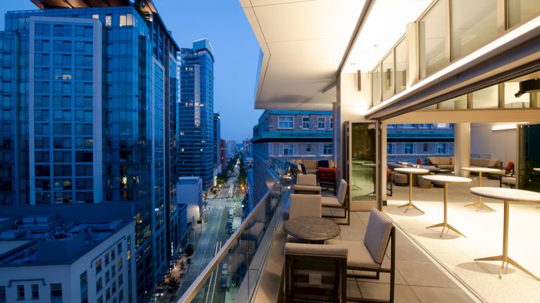 An outdoor balcony with seating overlooks a cityscape at dusk, featuring tall buildings and a well-lit street. The sentence ends here.