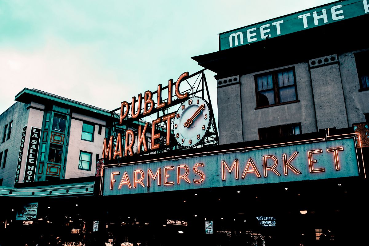 The image shows a neon sign for a "Farmers Market" at a "Public Market" with a clock above it, set against a backdrop of buildings.