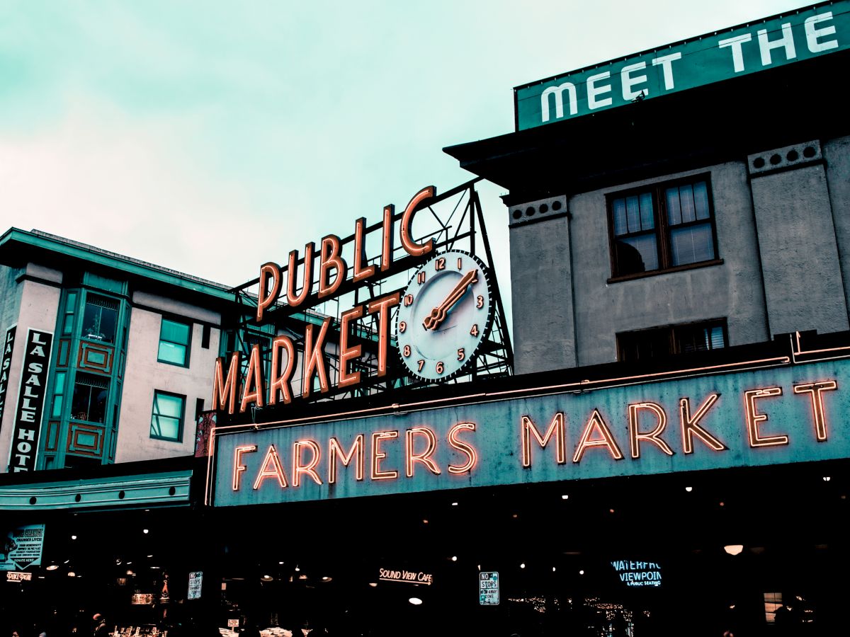 The image shows a neon sign for a "Farmers Market" at a "Public Market" with a clock above it, set against a backdrop of buildings.