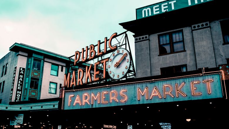 The image shows a neon sign for a "Farmers Market" at a "Public Market" with a clock above it, set against a backdrop of buildings.