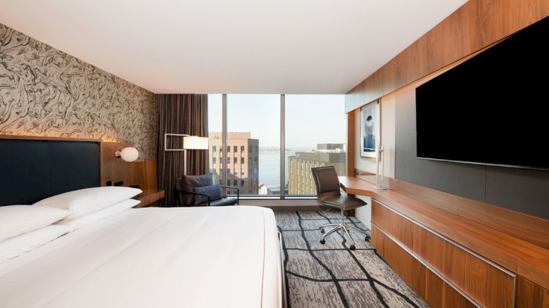 A modern hotel room features a large bed, flat-screen TV, desk, and floor-to-ceiling window with a city view, finished the sentence.