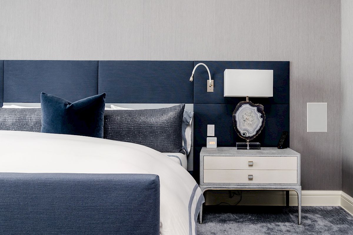 A modern bedroom featuring a bed with blue upholstery and throw pillows, a nightstand with a lamp, and a grey carpeted floor.