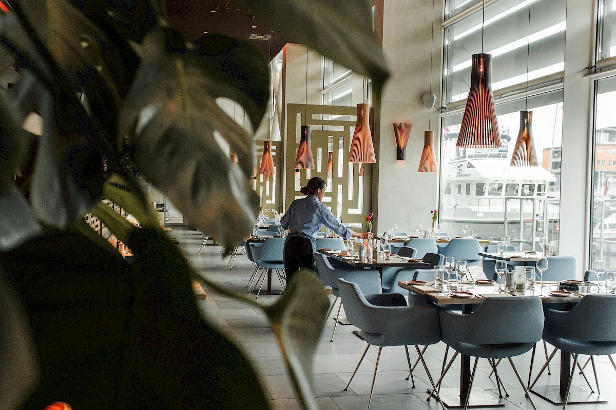 A waiter sets tables in a modern, stylish restaurant with large windows, plants, and unique pendant lighting.