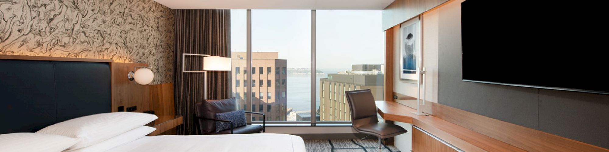 Modern hotel room with a large bed, wall-mounted TV, desk, and floor-to-ceiling windows revealing a cityscape view.