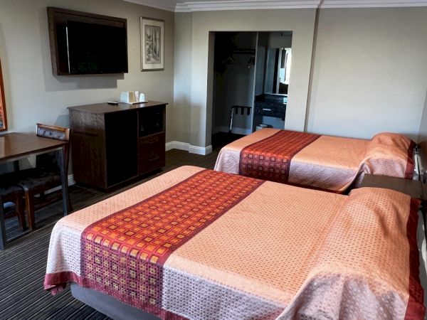 The image shows a hotel room with two beds, a TV, a small table with chairs, and a mirror on the wall. The decor is simple and neat.