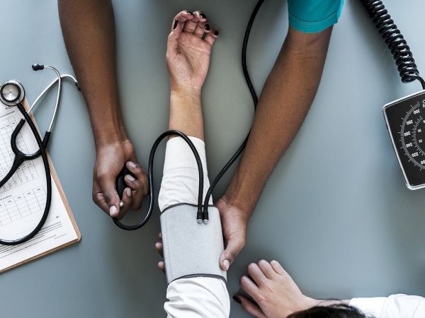 A healthcare professional is measuring a patient's blood pressure with a sphygmomanometer on a desk, with a stethoscope and clipboard nearby.